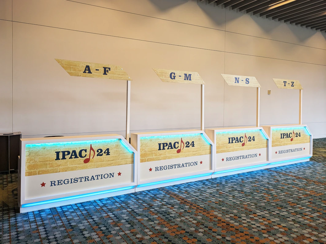 Four registration booths for the 15th International Particle Accelerator Conference, IPAC 24, are arranged side by side, each labeled with a different alphabet range: A-F, G-M, N-S, and T-Z. The white counters are lit with blue light and sit on a patterned carpet in a spacious indoor area. Large-format design by Phoenix Pheasant / Phoenix Pleasant.